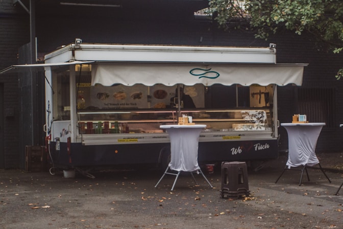 A food truck with a canopy is parked against a dark building. It is decorated with images of fish and sandwiches, suggesting it serves seafood. Two tall tables covered with white cloth are placed in front of the truck, each holding napkins and condiments. The area is littered with a few fallen leaves, and there is a tree partially visible to the right.
