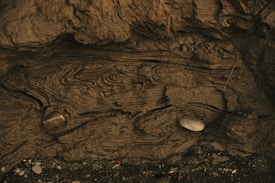 Intricate swirls and patterns appear on a rugged, textured surface resembling sedimentary rock or earth. Two smooth stones are embedded within the formations, one darker and larger, and the other lighter and smaller. The scene suggests natural erosion or weathering over time.