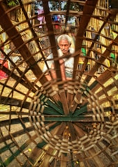 A man with grey hair is focusing intently on crafting a large basket, made from woven bamboo. He is seated in an outdoor setting surrounded by bamboo materials and other objects. The basket is the main foreground element, creating a circular frame around the craftsman.