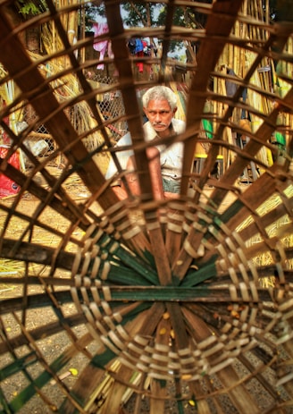 A man with grey hair is focusing intently on crafting a large basket, made from woven bamboo. He is seated in an outdoor setting surrounded by bamboo materials and other objects. The basket is the main foreground element, creating a circular frame around the craftsman.