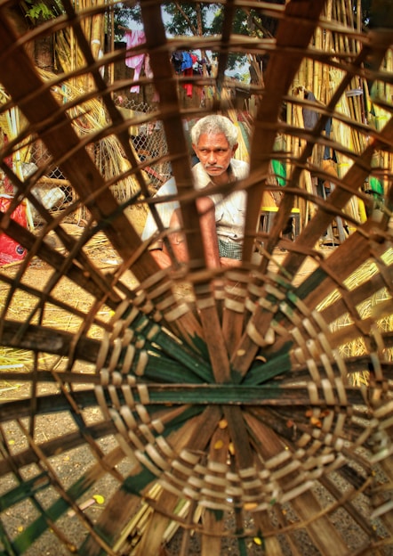 A man with grey hair is focusing intently on crafting a large basket, made from woven bamboo. He is seated in an outdoor setting surrounded by bamboo materials and other objects. The basket is the main foreground element, creating a circular frame around the craftsman.