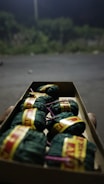 A close-up view of a cardboard box containing small cylindrical bundles wrapped in green cloth, secured with yellow and red labels, and tied with purple strings. The background is blurred, showing an outdoor setting with dim lighting and vegetation.