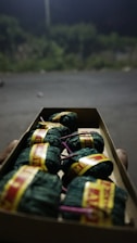 A close-up view of a cardboard box containing small cylindrical bundles wrapped in green cloth, secured with yellow and red labels, and tied with purple strings. The background is blurred, showing an outdoor setting with dim lighting and vegetation.