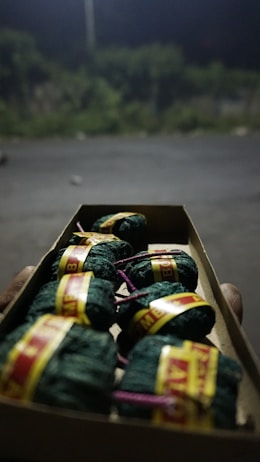 A close-up view of a cardboard box containing small cylindrical bundles wrapped in green cloth, secured with yellow and red labels, and tied with purple strings. The background is blurred, showing an outdoor setting with dim lighting and vegetation.