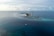 Aerial view of a coastal resort with overwater bungalows arranged in a circular pattern surrounded by clear blue ocean. Additional small islands are visible on the horizon, alongside several boats dotting the water. The sky is partly cloudy, adding to the serene and tropical setting.
