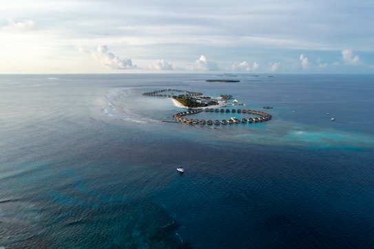 Aerial view of a coastal resort with overwater bungalows arranged in a circular pattern surrounded by clear blue ocean. Additional small islands are visible on the horizon, alongside several boats dotting the water. The sky is partly cloudy, adding to the serene and tropical setting.