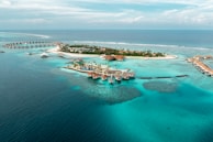 An aerial view of a tropical island resort with overwater bungalows extending into the turquoise waters of the ocean. The island is dotted with palm trees and surrounded by clear blue seas, with small areas of coral visible beneath the surface. There is a symmetrical arrangement of facilities and paths on the island, and the horizon shows a calm, expansive ocean meeting the sky.