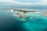 An aerial view of a tropical island resort with overwater bungalows extending into the turquoise waters of the ocean. The island is dotted with palm trees and surrounded by clear blue seas, with small areas of coral visible beneath the surface. There is a symmetrical arrangement of facilities and paths on the island, and the horizon shows a calm, expansive ocean meeting the sky.