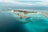An aerial view of a tropical island resort with overwater bungalows extending into the turquoise waters of the ocean. The island is dotted with palm trees and surrounded by clear blue seas, with small areas of coral visible beneath the surface. There is a symmetrical arrangement of facilities and paths on the island, and the horizon shows a calm, expansive ocean meeting the sky.