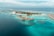 An aerial view of a tropical island resort with overwater bungalows extending into the turquoise waters of the ocean. The island is dotted with palm trees and surrounded by clear blue seas, with small areas of coral visible beneath the surface. There is a symmetrical arrangement of facilities and paths on the island, and the horizon shows a calm, expansive ocean meeting the sky.