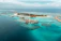 An aerial view of a tropical island resort with overwater bungalows extending into the turquoise waters of the ocean. The island is dotted with palm trees and surrounded by clear blue seas, with small areas of coral visible beneath the surface. There is a symmetrical arrangement of facilities and paths on the island, and the horizon shows a calm, expansive ocean meeting the sky.