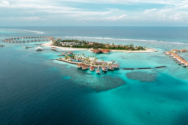 An aerial view of a tropical island resort with overwater bungalows extending into the turquoise waters of the ocean. The island is dotted with palm trees and surrounded by clear blue seas, with small areas of coral visible beneath the surface. There is a symmetrical arrangement of facilities and paths on the island, and the horizon shows a calm, expansive ocean meeting the sky.