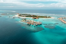 An aerial view of a tropical island resort with overwater bungalows extending into the turquoise waters of the ocean. The island is dotted with palm trees and surrounded by clear blue seas, with small areas of coral visible beneath the surface. There is a symmetrical arrangement of facilities and paths on the island, and the horizon shows a calm, expansive ocean meeting the sky.