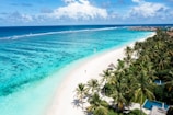 Aerial view of a tropical beach with white sand, surrounded by lush green palm trees and clear turquoise water stretching into the distance. Overwater bungalows line the horizon on the right, while a single person walks along the shoreline.