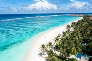 Aerial view of a tropical beach with white sand, surrounded by lush green palm trees and clear turquoise water stretching into the distance. Overwater bungalows line the horizon on the right, while a single person walks along the shoreline.
