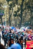 A vibrant parade with people wearing colorful costumes and masks, taking place on a street lined with tall trees. Many participants carry umbrellas, and some wear skeleton-themed outfits. The scene is bustling with spectators, some of whom are taking photos. Signs visible in the background provide a sense of location.