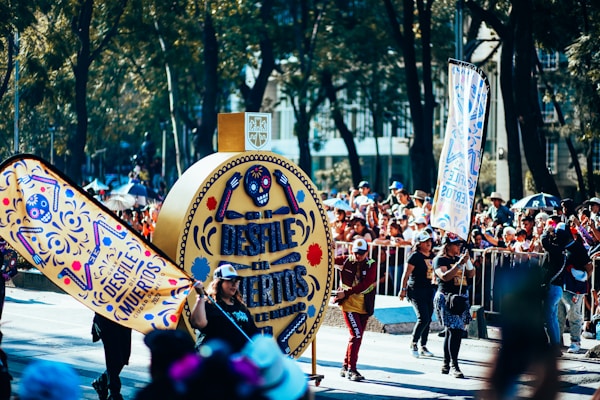 A vibrant parade with numerous participants and onlookers. The central focus is on an intricately designed float bearing the text 'Desfile de Muertos'. People are dressed in festive costumes, some holding decorative banners matching the float. The background is filled with trees and buildings while the street is lined with metal barriers holding back the crowd.