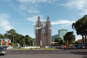 A large structure is under renovation or construction, covered in scaffolding, situated in an urban area. The surrounding environment includes a traffic circle with vehicles, pedestrians, and modern buildings, including one labeled Diamond Plaza. The scene is framed by trees and a clear blue sky.