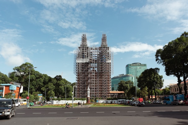A large structure is under renovation or construction, covered in scaffolding, situated in an urban area. The surrounding environment includes a traffic circle with vehicles, pedestrians, and modern buildings, including one labeled Diamond Plaza. The scene is framed by trees and a clear blue sky.