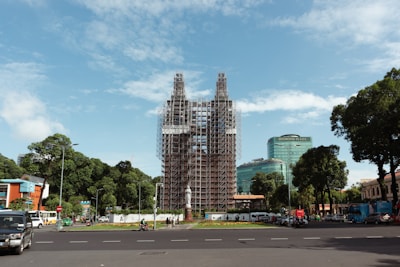 A large structure is under renovation or construction, covered in scaffolding, situated in an urban area. The surrounding environment includes a traffic circle with vehicles, pedestrians, and modern buildings, including one labeled Diamond Plaza. The scene is framed by trees and a clear blue sky.
