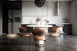 A modern kitchen interior with sleek, dark cabinetry and a minimalist design. The room features a large refrigerator on the left, built-in cabinetry around a sink and countertop, and a stylish round dining table with four chairs. Above the table hangs a distinctive black pendant light fixture. On the countertop are a coffee maker, decorative vases, and potted plants, adding an aesthetic touch.