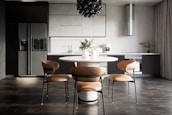 A modern kitchen interior with sleek, dark cabinetry and a minimalist design. The room features a large refrigerator on the left, built-in cabinetry around a sink and countertop, and a stylish round dining table with four chairs. Above the table hangs a distinctive black pendant light fixture. On the countertop are a coffee maker, decorative vases, and potted plants, adding an aesthetic touch.