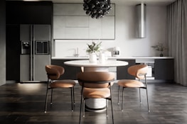 A modern kitchen interior with sleek, dark cabinetry and a minimalist design. The room features a large refrigerator on the left, built-in cabinetry around a sink and countertop, and a stylish round dining table with four chairs. Above the table hangs a distinctive black pendant light fixture. On the countertop are a coffee maker, decorative vases, and potted plants, adding an aesthetic touch.
