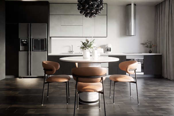 A modern kitchen interior with sleek, dark cabinetry and a minimalist design. The room features a large refrigerator on the left, built-in cabinetry around a sink and countertop, and a stylish round dining table with four chairs. Above the table hangs a distinctive black pendant light fixture. On the countertop are a coffee maker, decorative vases, and potted plants, adding an aesthetic touch.