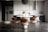 A modern kitchen interior with sleek, dark cabinetry and a minimalist design. The room features a large refrigerator on the left, built-in cabinetry around a sink and countertop, and a stylish round dining table with four chairs. Above the table hangs a distinctive black pendant light fixture. On the countertop are a coffee maker, decorative vases, and potted plants, adding an aesthetic touch.