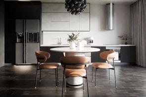 A modern kitchen interior with sleek, dark cabinetry and a minimalist design. The room features a large refrigerator on the left, built-in cabinetry around a sink and countertop, and a stylish round dining table with four chairs. Above the table hangs a distinctive black pendant light fixture. On the countertop are a coffee maker, decorative vases, and potted plants, adding an aesthetic touch.