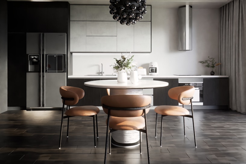A modern kitchen interior with sleek, dark cabinetry and a minimalist design. The room features a large refrigerator on the left, built-in cabinetry around a sink and countertop, and a stylish round dining table with four chairs. Above the table hangs a distinctive black pendant light fixture. On the countertop are a coffee maker, decorative vases, and potted plants, adding an aesthetic touch.