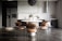 A modern kitchen interior with sleek, dark cabinetry and a minimalist design. The room features a large refrigerator on the left, built-in cabinetry around a sink and countertop, and a stylish round dining table with four chairs. Above the table hangs a distinctive black pendant light fixture. On the countertop are a coffee maker, decorative vases, and potted plants, adding an aesthetic touch.