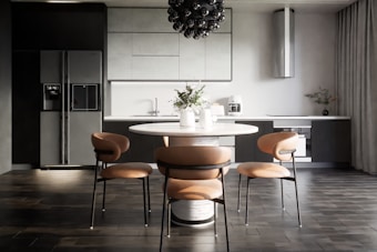 A modern kitchen interior with sleek, dark cabinetry and a minimalist design. The room features a large refrigerator on the left, built-in cabinetry around a sink and countertop, and a stylish round dining table with four chairs. Above the table hangs a distinctive black pendant light fixture. On the countertop are a coffee maker, decorative vases, and potted plants, adding an aesthetic touch.