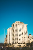 A tall residential building with numerous windows and balconies stands against a clear blue sky. Surrounding the building are palm trees and other urban features such as street lights and traffic signs. The architecture includes multiple floors with a modern design.