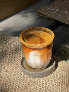 A glass of coffee sits on a round stone coaster, placed on a woven fabric surface. The coffee has a rich, creamy swirl pattern with a frothy top and a decorative sugar rim around the glass.