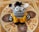 A knitted stuffed toy with large, expressive black and white eyes, black and white striped pattern, adorned with a tuft of black yarn on top of its head. It wears a vibrant yellow crochet outfit fastened with two yellow buttons, set against an intricately patterned tile background with ornate designs.