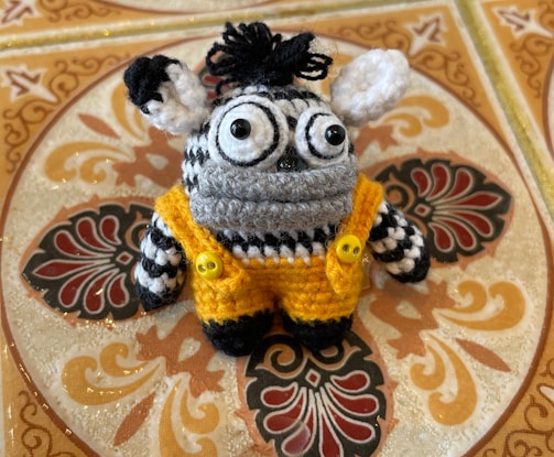 A knitted stuffed toy with large, expressive black and white eyes, black and white striped pattern, adorned with a tuft of black yarn on top of its head. It wears a vibrant yellow crochet outfit fastened with two yellow buttons, set against an intricately patterned tile background with ornate designs.