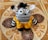 A knitted stuffed toy with large, expressive black and white eyes, black and white striped pattern, adorned with a tuft of black yarn on top of its head. It wears a vibrant yellow crochet outfit fastened with two yellow buttons, set against an intricately patterned tile background with ornate designs.