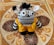 A knitted stuffed toy with large, expressive black and white eyes, black and white striped pattern, adorned with a tuft of black yarn on top of its head. It wears a vibrant yellow crochet outfit fastened with two yellow buttons, set against an intricately patterned tile background with ornate designs.