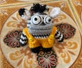 A knitted stuffed toy with large, expressive black and white eyes, black and white striped pattern, adorned with a tuft of black yarn on top of its head. It wears a vibrant yellow crochet outfit fastened with two yellow buttons, set against an intricately patterned tile background with ornate designs.