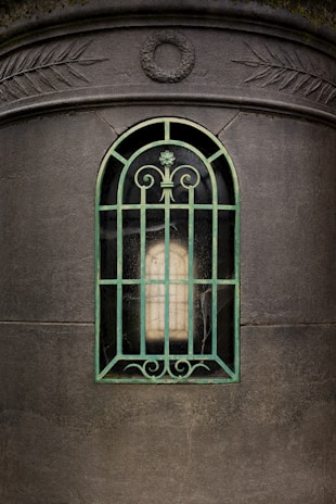 An arched window with a decorative metal grill in front of a stone structure. The metal grill is painted green and has intricate scrollwork at the top and bottom. The background within the window appears dark and old, giving a mysterious and ancient feel. On the stone structure above the window, there are carved patterns of leaves and a circular design in the center.