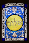 A stained glass window featuring elaborate floral patterns in blue and white surrounding a central yellow circle. Inside the circle, Hebrew text is prominently displayed. The design exhibits detailed craftsmanship with symmetrical motifs.
