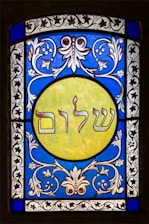 A stained glass window featuring elaborate floral patterns in blue and white surrounding a central yellow circle. Inside the circle, Hebrew text is prominently displayed. The design exhibits detailed craftsmanship with symmetrical motifs.