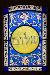 A stained glass window featuring elaborate floral patterns in blue and white surrounding a central yellow circle. Inside the circle, Hebrew text is prominently displayed. The design exhibits detailed craftsmanship with symmetrical motifs.
