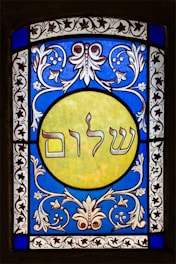 A stained glass window featuring elaborate floral patterns in blue and white surrounding a central yellow circle. Inside the circle, Hebrew text is prominently displayed. The design exhibits detailed craftsmanship with symmetrical motifs.