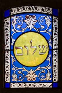 A stained glass window featuring elaborate floral patterns in blue and white surrounding a central yellow circle. Inside the circle, Hebrew text is prominently displayed. The design exhibits detailed craftsmanship with symmetrical motifs.