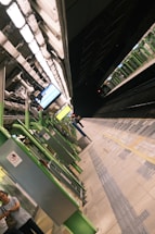 A train station platform with green and white structural elements. The platform is empty except for a couple of people in the foreground, and a train is visible on the track in the background. Overhead lighting illuminates the area, and digital signs are present.