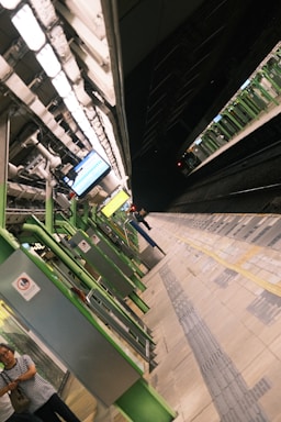 A train station platform with green and white structural elements. The platform is empty except for a couple of people in the foreground, and a train is visible on the track in the background. Overhead lighting illuminates the area, and digital signs are present.