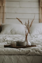 A cozy and rustic bedroom scene with a neatly made bed covered in soft, beige bedding. On the bed, a wooden tray holds a transparent vase containing dried grasses and two ceramic mugs, creating a warm and inviting atmosphere.