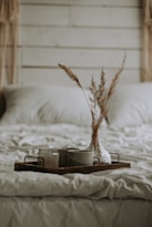A cozy and rustic bedroom scene with a neatly made bed covered in soft, beige bedding. On the bed, a wooden tray holds a transparent vase containing dried grasses and two ceramic mugs, creating a warm and inviting atmosphere.